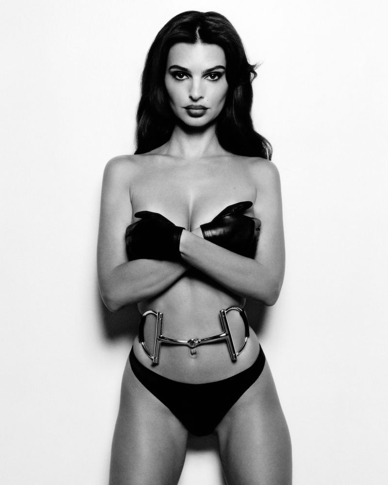 Black-and-white studio shot of a woman wearing a black thong and gloves with a silver metal horse bit belt, striking a confident pose.