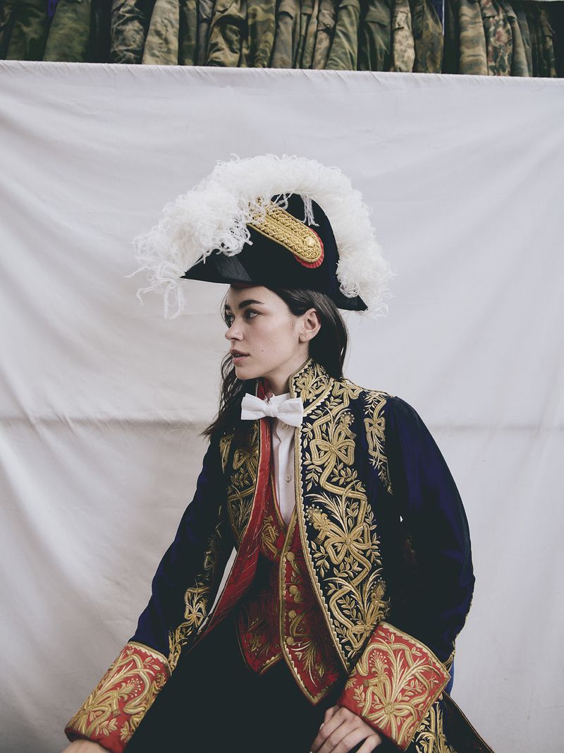 Woman in a historical-inspired ensemble featuring an ornate blue coat with gold detailing, a white bow tie, and a feathered tricorn hat.