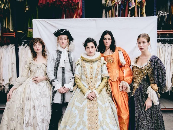 Five women pose in a row, wearing elaborate renaissance-style gowns in varied colors with a costume rack and white sheet behind them.