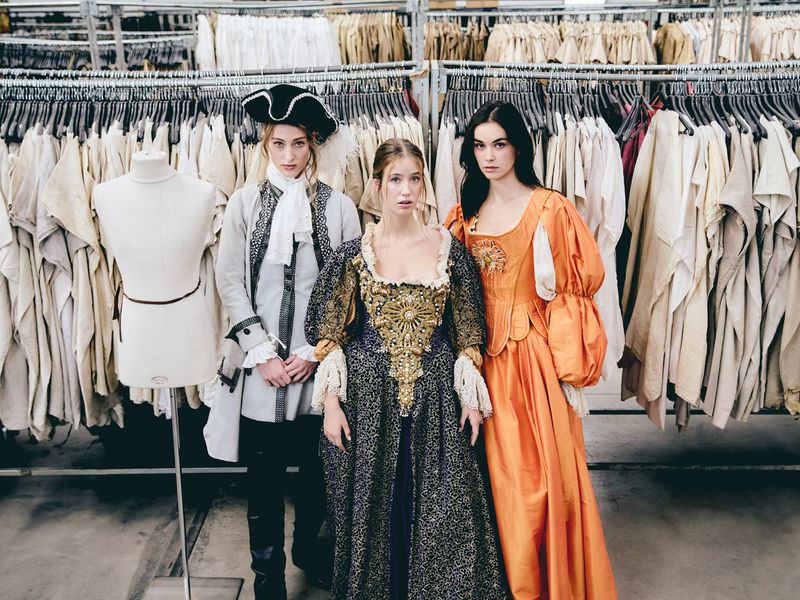 Three women in historical costumes posing in front of a rack of clothes, indoor setting with neutral colors and studio lighting, fashion concept.