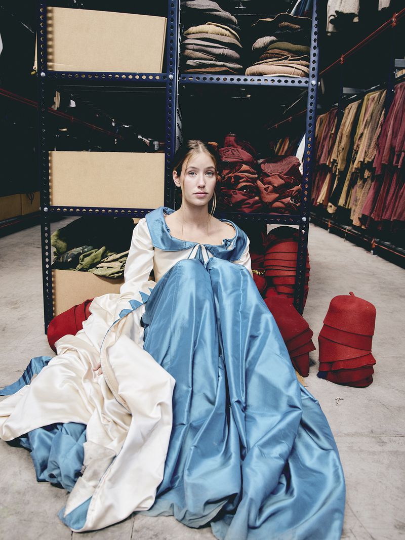 Young woman in elaborate blue gown sits on floor in costume warehouse surrounded by garments on shelves. Indoor fashion portrait.