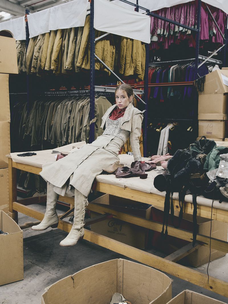 Young woman in beige coat and high boots poses on table filled with clothing and shoes in large warehouse setting.