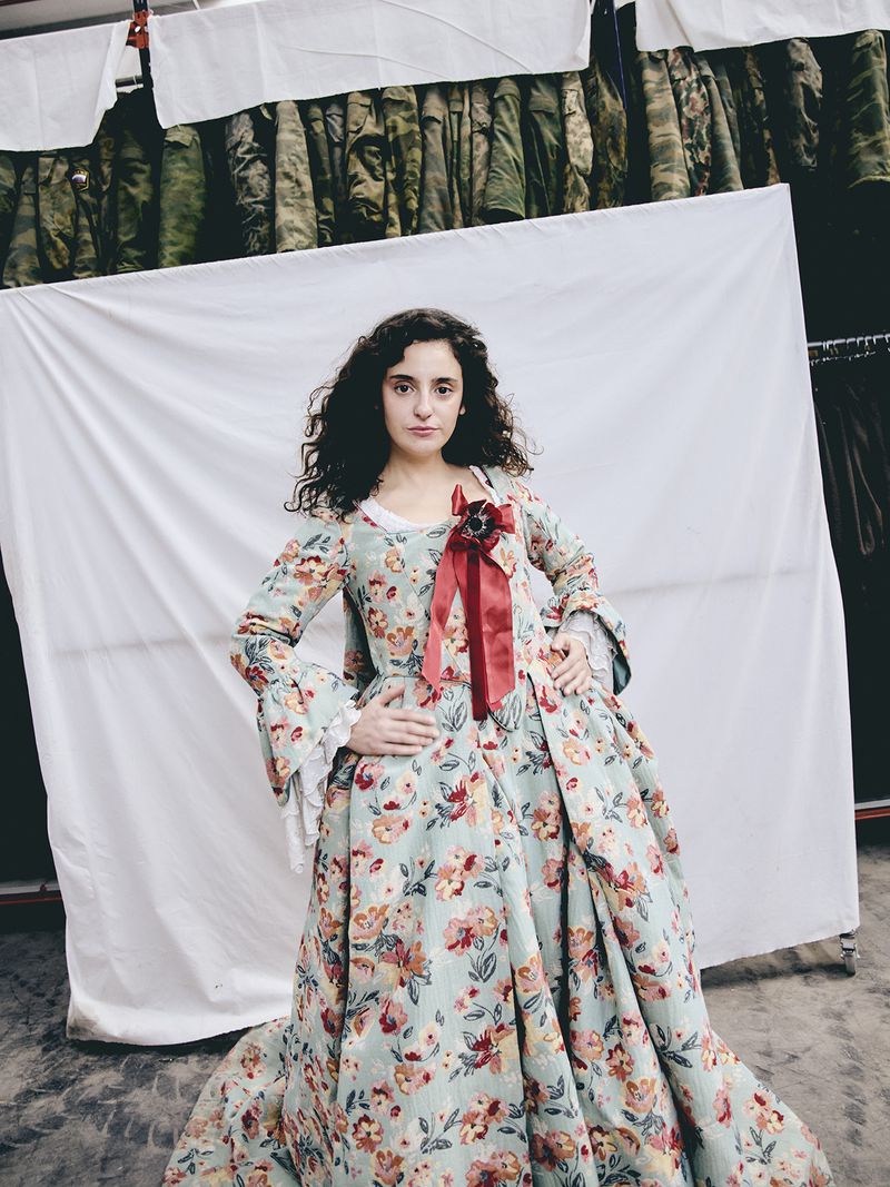 A curly-haired woman poses in a light blue floral gown with a red ribbon against a backdrop of white fabric and draped green garments.