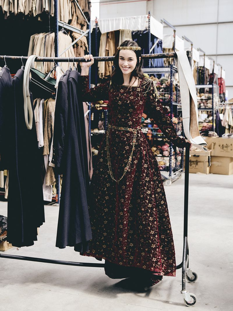 A smiling woman in a medieval-style dress stands in a warehouse filled with clothing racks and costumes in a medium, full shot.