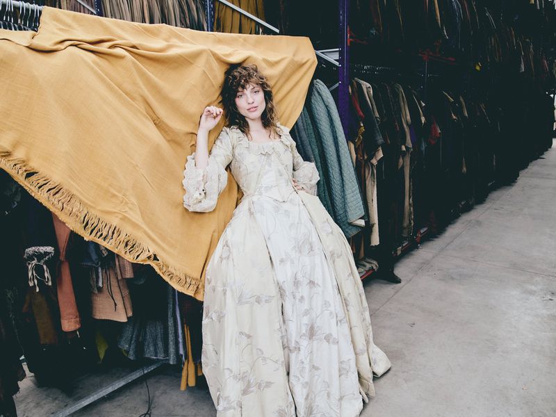Woman in vintage dress lies under yellow textile in warehouse. Model poses on fabric rack with period gown in a large indoor space.