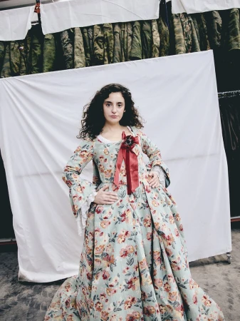 Woman wearing an 18th-century style floral gown with a red chest ribbon standing before a draped white sheet backdrop.