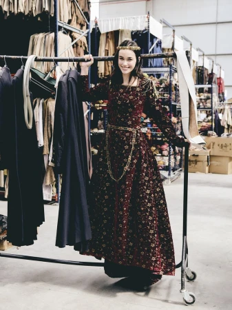 Smiling woman wearing a long patterned burgundy dress holding onto a clothing rack in a warehouse setting.