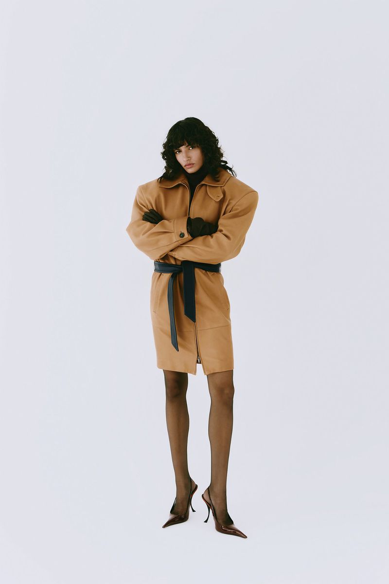 A woman in a tan belted coat and black slingback heels stands in a white studio, arms crossed, radiating confidence and style.