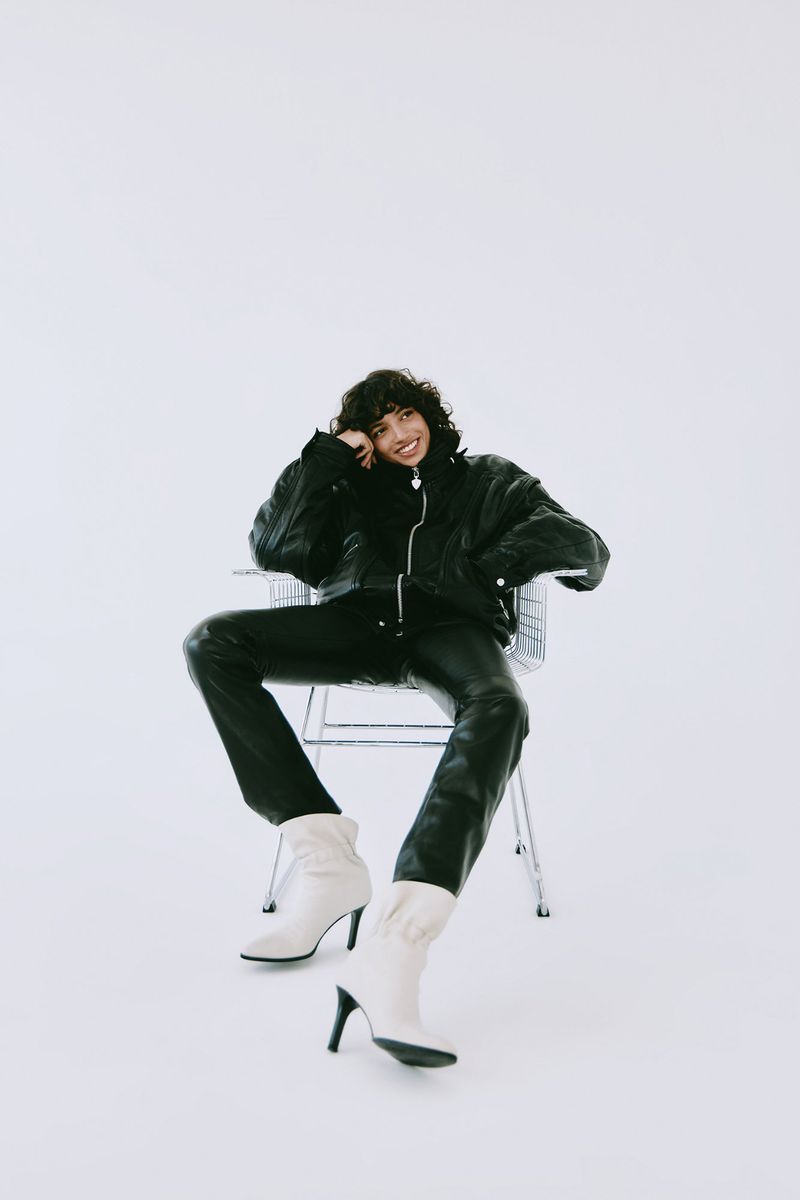 Fashionable woman in a black leather jacket and pants, seated on a silver chair in a bright white studio, wearing white heeled boots.