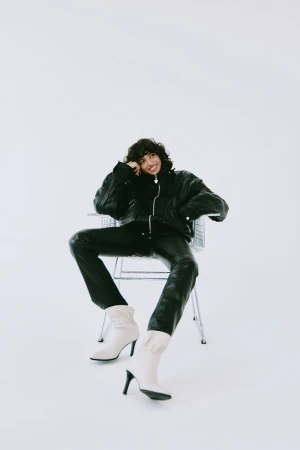 Fashionable woman in a black leather jacket and pants, seated on a silver chair in a bright white studio, wearing white heeled boots.