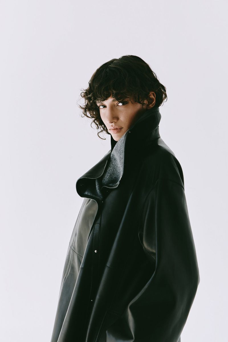 Model with dark curly hair wearing a black leather coat with a high collar against a plain white background.