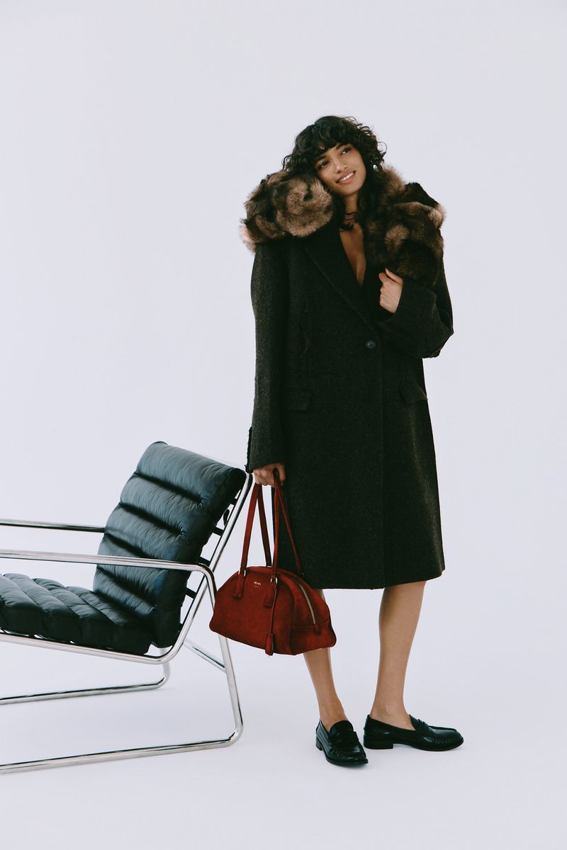 Stylish woman in a fur-trimmed coat stands in a bright studio, holding a red leather bag, with a black leather chair nearby.