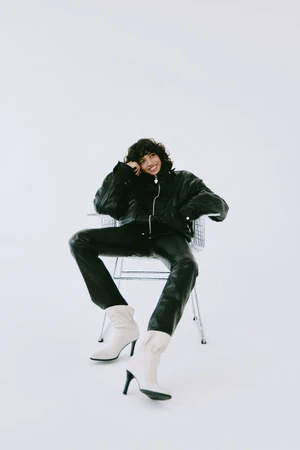 Model Maria Khan smiling while seated in a metal wire chair wearing a black jacket, leather pants, and cream high-heeled boots.