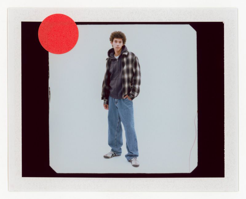 A young adult male stands indoors against a gray background, wearing jeans, a grey hooded top and a checkered jacket, in a fashion shot.
