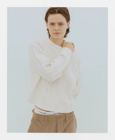 Portrait of a young adult female in a white knit sweater and brown corduroy pants against a light blue background in a photo studio.