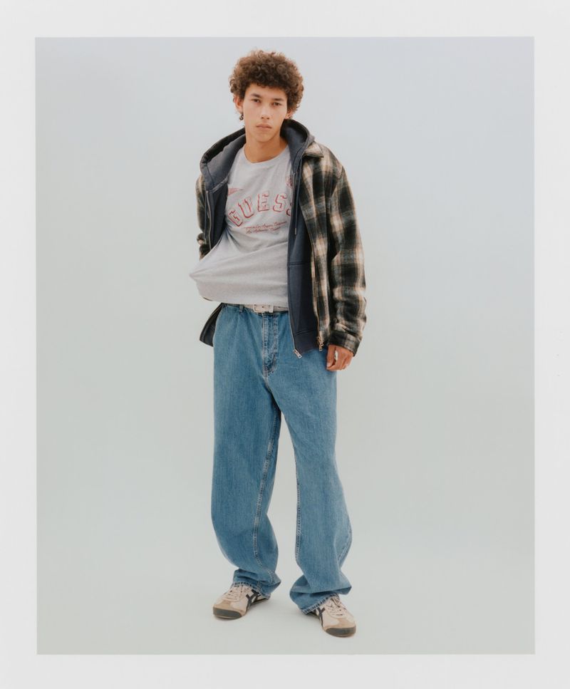 Young man in a gray branded T-shirt, flannel, and jeans, standing in a studio with neutral lighting, creating a casual, stylish portrait.