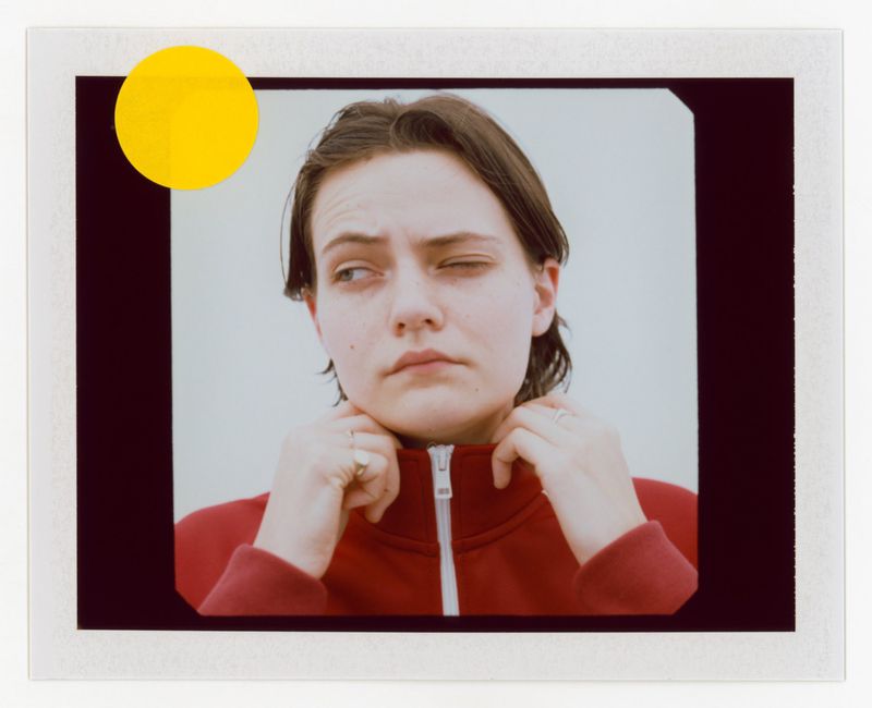 Young woman with short brown hair poses indoors in a red zip-up jacket against a light background, with a bright yellow sticker.