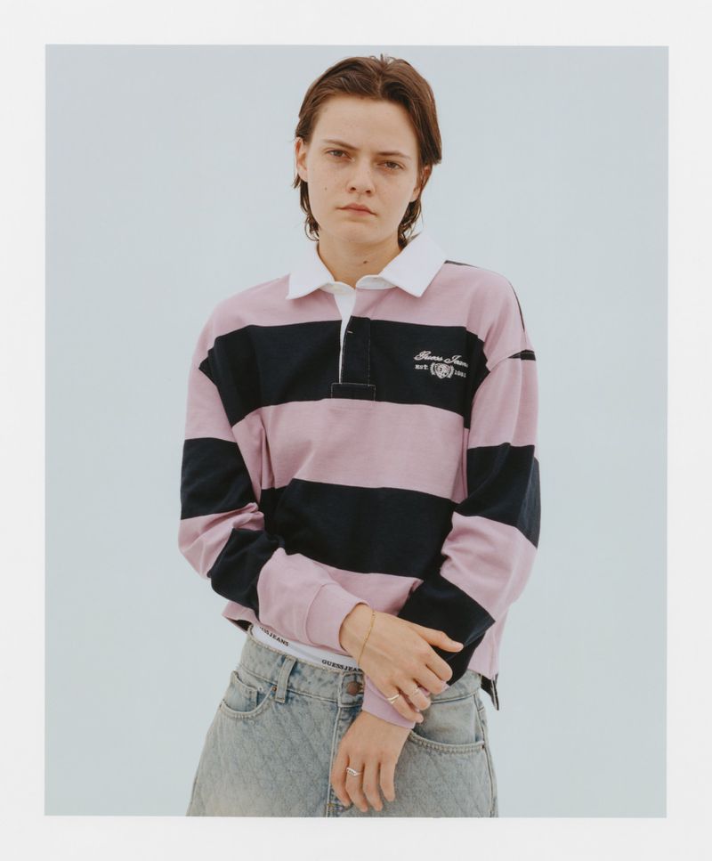 Young adult woman in a pink and black striped rugby shirt with light wash jeans posing in a bright indoor studio setting. 
