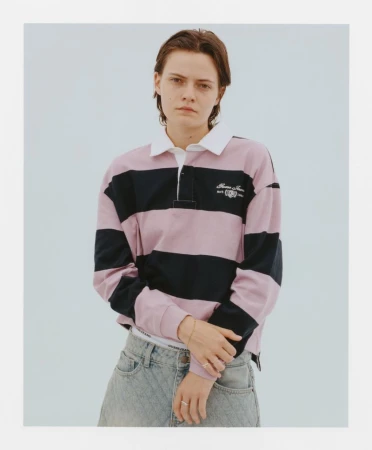 Young adult woman in a pink and black striped rugby shirt with light wash jeans posing in a bright indoor studio setting. 