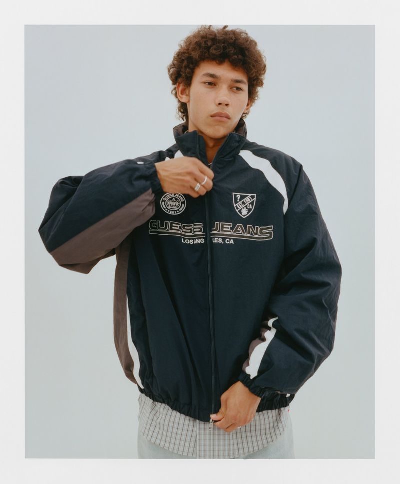 Young adult man in a dark blue Guess Jeans track jacket with white and grey accents, standing in a neutral studio setting.