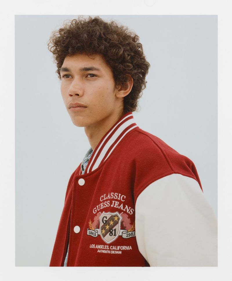 A young man with curly hair poses in a studio wearing a red and white GUESS varsity jacket against a soft blue-grey backdrop for a fashion editorial.