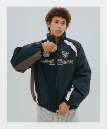 Young man wearing a dark nylon zip jacket with white paneling and Guess Jeans branding against a neutral grey studio background.
