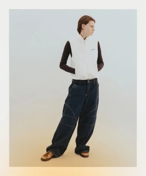 Model wearing a white puffer vest and dark denim jeans standing against a plain gradient background.