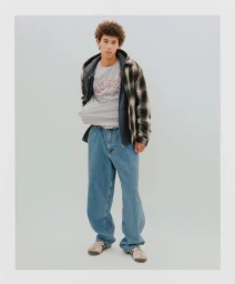 Young man with curly hair wearing a plaid jacket over grey t-shirt and loose blue jeans against a light grey background