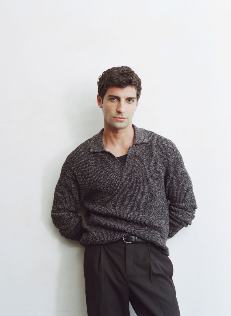 Portrait of an adult male with dark hair, wearing a grey knit sweater and black pants, posing against a white wall indoors.