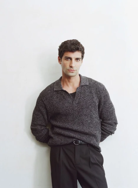 Portrait of an adult male with dark hair, wearing a grey knit sweater and black pants, posing against a white wall indoors.
