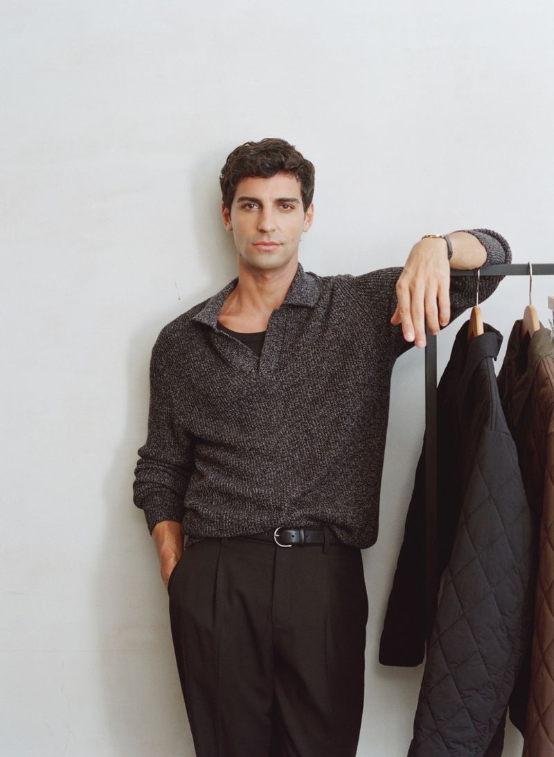 An adult male poses indoors in a grey polo shirt and black slacks, leaning on a rack of dark jackets against a neutral backdrop.