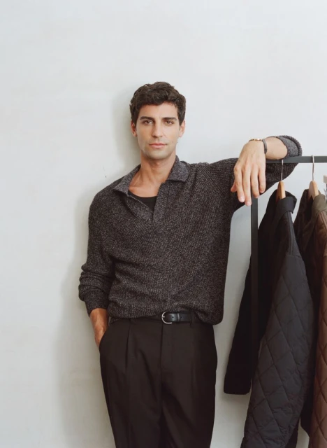 An adult male poses indoors in a grey polo shirt and black slacks, leaning on a rack of dark jackets against a neutral backdrop.