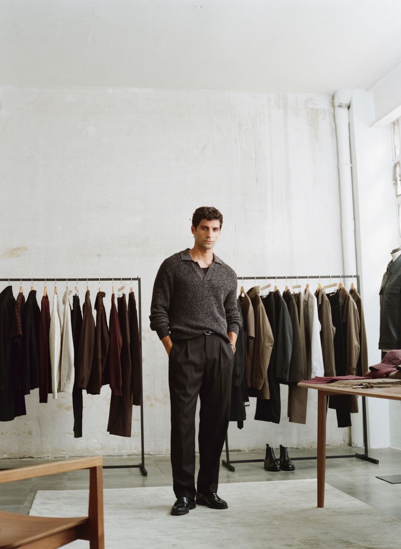 A fashionable adult male poses indoors, dressed in a grey sweater and dark trousers, with clothing racks in the background.