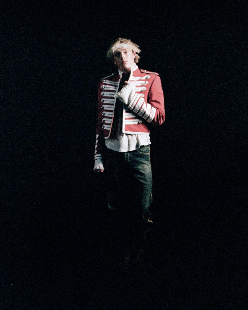 Solo adult male with blond hair in a red and white military-style jacket and jeans, standing in a dark studio against a black background.