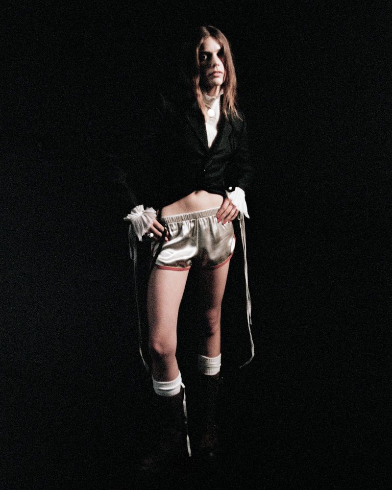 A woman stands in a dark space modeling a black blazer with billowing white sleeves and silver shorts with red trim and knee high socks.