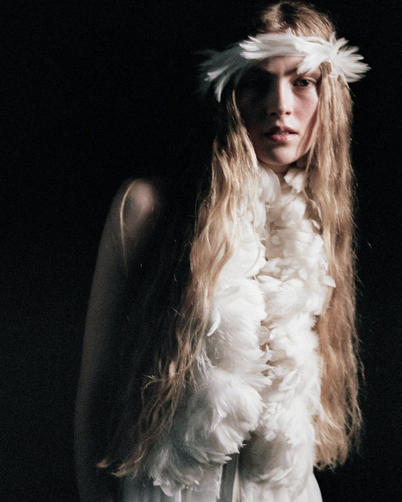 A woman with long blonde hair poses in a white feather boa and feathered headband against a dark background in a studio setting.