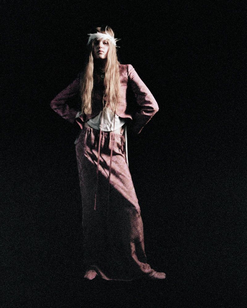 A blonde model in a mauve outfit poses against a dark background, wearing a feather headdress and an unbuttoned jacket over a white top.