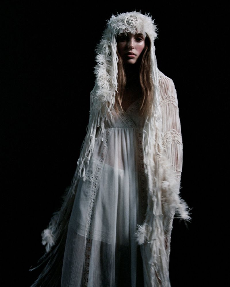 Fashionable woman with feathered headpiece and gown posing in a dark studio, evoking an ethereal and high-fashion aesthetic.