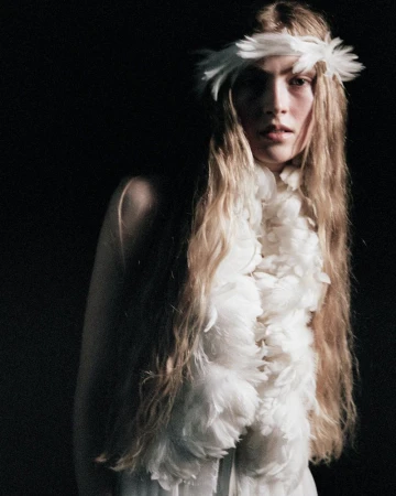 Model wearing a white feathered headdress and feather scarf against a dark background for Ann Demeulemeester