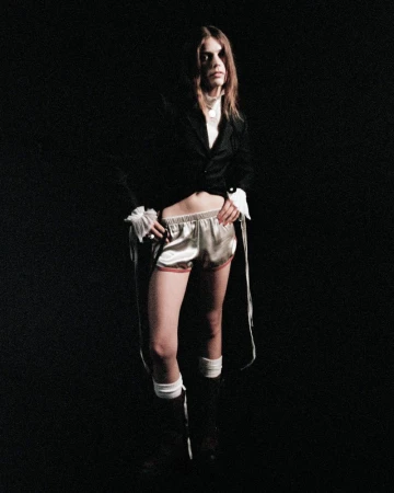 Model wearing a black jacket and satin shorts against a dark background for Ann Demeulemeester