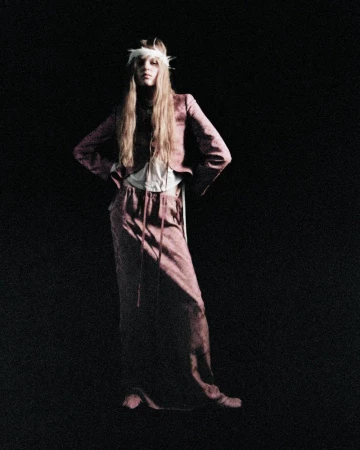 Model standing against a dark background wearing a pink patterned blazer and long skirt with a feather headband.