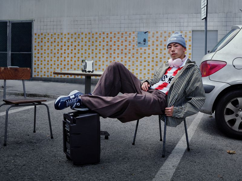 Asian man in streetwear fashion poses on a chair in an urban parking lot next to a silver car, wearing headphones and a beanie.