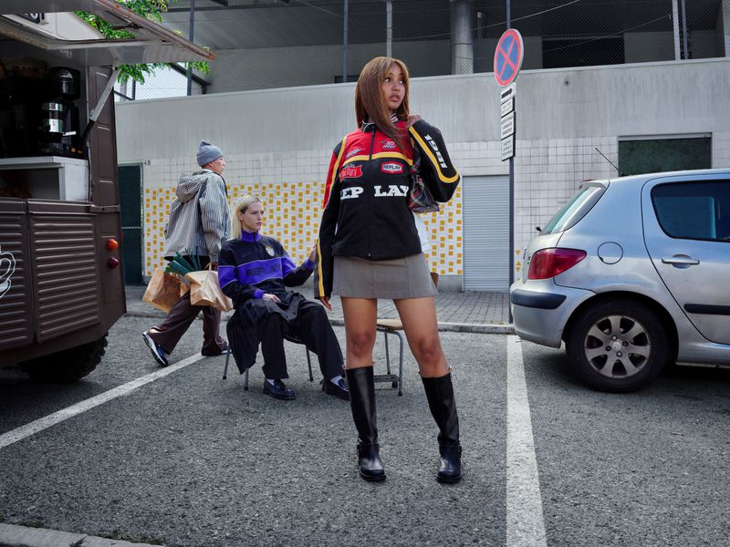 Young woman in racing jacket and mini skirt poses near food truck and parked car with two other figures in the background.