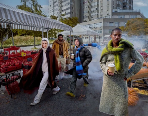 Fashion editorial shot outdoors at a farmer's market with four diverse adults wearing contemporary knitwear and outerwear, holding coffee.