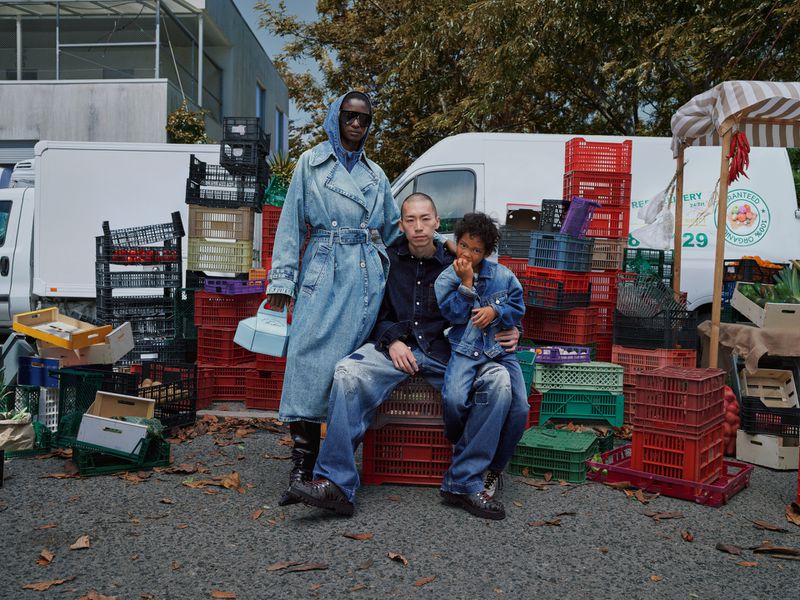 Three people posing with a backdrop of stacked crates, including a white van and a fruit stand, outdoors in daytime with natural lighting.