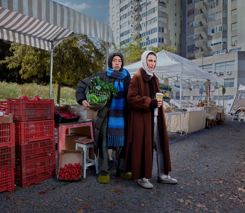 Two adults stand at a market, one holding a basket of vegetables, the other a coffee cup, with produce stands and city buildings visible.