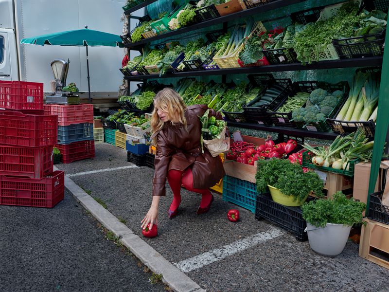 Woman in brown jacket and red tights crouching to pick up a red bell pepper at outdoor market with produce on display.