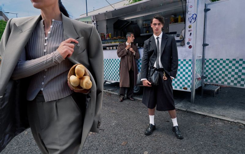 Three people stand outside a food truck. A person in a grey suit holds a bowl of bread rolls in the foreground. Editorial fashion.