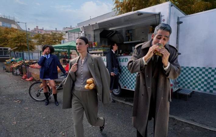Fashion shot of people in business attire near a food truck and produce stand, featuring a woman carrying bread, and a man eating a sandwich.