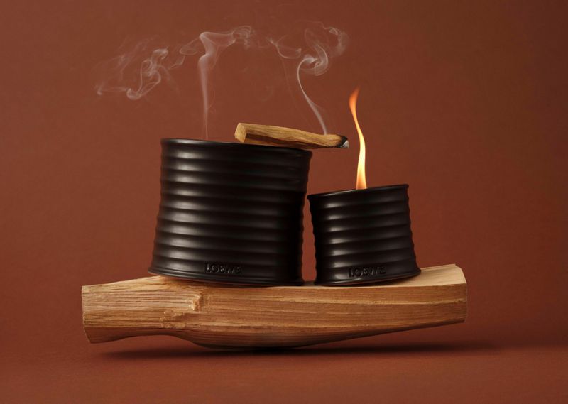 Two ceramic candle holders on a wooden block, one with a flame and palo santo incense stick emitting smoke against brown backdrop.